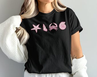 Może przedstawiać: Czarny t-shirt z różowymi grafikami o tematyce morskiej: rozgwiazda, muszla, krab i muszla spiralna. Projekt jest wyśrodkowany na klatce piersiowej. Koszulka jest codziennym, swobodnym elementem garderoby.
