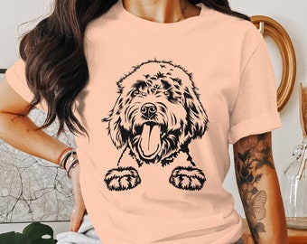 Puede incluir: Camiseta color melocotón con un gráfico negro de la cara de un perro con la lengua fuera. El pelaje del perro está detallado con líneas intrincadas, y el diseño está centrado en la camiseta.