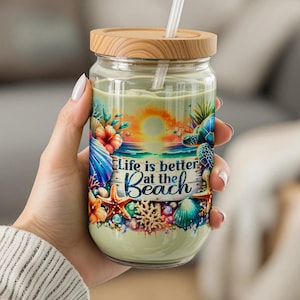 May include: A glass tumbler with a wooden lid and straw, featuring a beach-themed design. The tumbler is decorated with seashells, starfish, and a sunset, with the text "Life is better at the Beach."