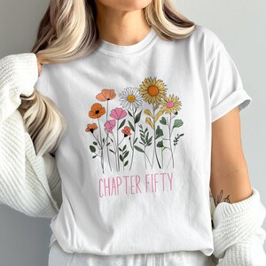 May include: White t-shirt featuring a floral design with orange, pink, yellow, and white flowers. The text "CHAPTER FIFTY" is printed in pink below the flowers. The shirt is a casual, everyday wear item.