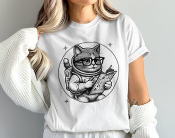 May include: White t-shirt featuring a black and white illustration of a cat astronaut wearing glasses and holding a clipboard. The cat is inside a circular frame with stars. The t-shirt is a casual, unisex style.