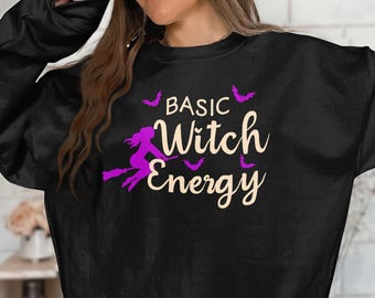 以下が含まれることがあります： 「BASIC Witch Energy」の文字が書かれた黒いスウェットシャツ。紫色のほうきに乗った魔女と紫色のコウモリのシルエットが描かれています。デザインは中央に配置されています。
