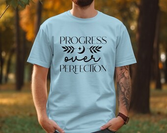 May include: A light blue short-sleeved t-shirt featuring the black text "PROGRESS over PERFECTION" centred on the chest. The design includes a small crescent moon and star icon flanked by two leafy branch illustrations between the lines of text.