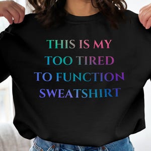 May include: Black sweatshirt with the text "THIS IS MY TOO TIRED TO FUNCTION SWEATSHIRT" in a gradient of colors. The text transitions from green to purple. The sweatshirt is a crew neck style.