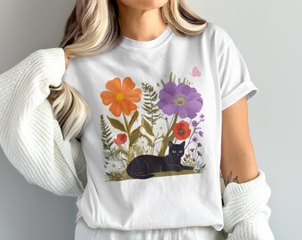 May include: White t-shirt featuring a black cat resting among colorful flowers. The design includes orange, purple, and red flowers, green foliage, and a pink butterfly. The cat has blue eyes.