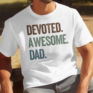 May include: White t-shirt with the words "DEVOTED. AWESOME. DAD." in brown, teal, and blue lettering. The shirt is a crew neck and is being worn by a person.
