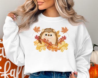 May include: White sweatshirt featuring a smiling hedgehog surrounded by colorful autumn leaves. The hedgehog has brown fur and a light beige face. The leaves are in shades of red, orange, and yellow, creating a fall theme.