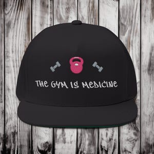Puede incluir: Gorra negra de ala plana con parte inferior verde. La gorra presenta bordados con temática de fitness: una pesa rusa rosa, dos mancuernas grises y el texto "THE GYM IS MEDICINE" en blanco.