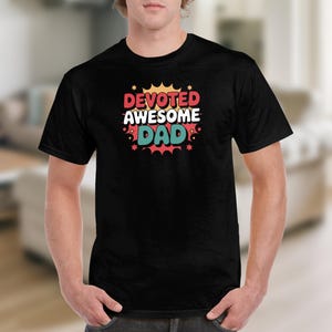 Peut inclure: T-shirt noir avec l'inscription "DEVOTED AWESOME DAD" dans une police colorée de style bande dessinée. Le texte est en rouge, blanc et sarcelle, avec des accents d'éclat d'étoile. Le t-shirt est porté par une personne.
