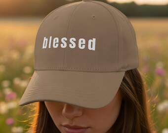 Może przedstawiać: Beżowa czapka z daszkiem z białym napisem "blessed". Czapka ma zakrzywiony daszek i zaokrągloną koronę. Tło to rozmyte pole kwiatów.