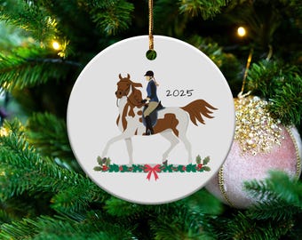 Könnte beinhalten: Ein weißer Keramik-Weihnachtsbaumschmuck mit einem braunen und weißen Pferd und einem Reiter in einer blauen Jacke. Das Pferd trägt einen braunen Sattel und der Reiter hält die Zügel. Der Schmuck hat einen grünen Stechpalmenkranz mit roten Beeren und einer roten Schleife unten. Das Jahr 2025 ist in Schwarz auf dem Schmuck geschrieben.
