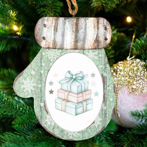 May include: A mitten-shaped Christmas ornament featuring a rustic corrugated metal cuff and a light green background with a white snowflake pattern. The center displays an oval illustration of a stack of three wrapped gift boxes with bows.