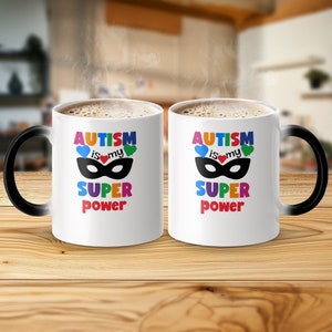 May include: Two white ceramic mugs with black handles, each displaying the text "AUTISM is my SUPER POWER" in colorful letters. The design includes a black mask, blue and green hearts, and a red heart.