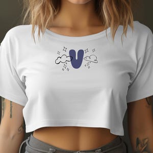 May include: White crop top with a blue letter "U" design, clouds, and stars. The shirt is a casual style, suitable for everyday wear.