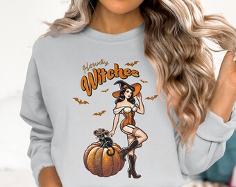 Może przedstawiać: Jasnoszara bluza z grafiką o tematyce Halloween. Projekt zawiera wiedźmę w stylu pin-up z czarnym kotem na dyni, nietoperze i napis "Howdy Witches" w kolorze pomarańczowym i brązowym.