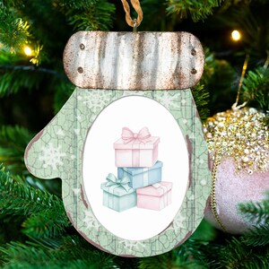May include: A mitten-shaped hanging ornament featuring a weathered metal cuff and a light green background with white snowflake patterns. The center displays an oval illustration of a stack of four gift boxes in pastel pink and light blue.