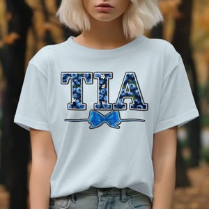 Op de afbeelding: Lichtblauw T-shirt met het woord "TIA" in grote blokletters gevuld met bosbessen en kleine witte bloemen. Een blauwe strik is gecentreerd onder de letters, met een dunne lijn die de strik met de letters verbindt.