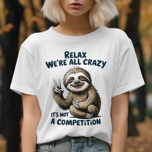 May include: White t-shirt with a cartoon sloth illustration. The sloth is making a peace sign and the shirt has the text "Relax We're All Crazy It's Not A Competition" in dark blue. The shirt is short-sleeved.