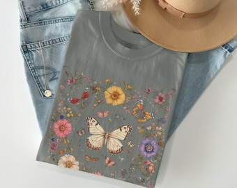 Peut inclure: Un t-shirt gris avec un motif floral et papillon. Le motif comprend un grand papillon blanc, des fleurs orange, roses et jaunes, et plusieurs petits papillons. Le t-shirt est plié sur un jean bleu et un chapeau beige.