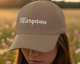 Puede incluir: Gorra de béisbol beige con el nombre "Maryann" bordado en escritura blanca. La gorra tiene una visera curva y un botón en la parte superior. El fondo es un campo de flores borroso.