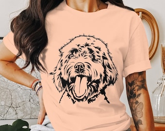 Puede incluir: Camiseta color melocotón con un gráfico negro de la cara de un perro con la lengua fuera. El pelaje del perro está detallado con líneas intrincadas, creando una apariencia texturizada. La camiseta tiene mangas cortas y un ajuste relajado, adecuada para uso casual.