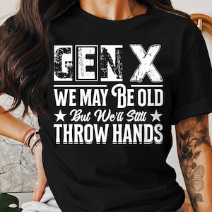 May include: Black t-shirt with white text reading "GEN X WE MAY BE OLD But We'll Still THROW HANDS". The text is in a distressed font. The shirt is a crew neck with short sleeves.