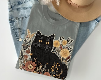 May include: A gray t-shirt featuring a black cat illustration surrounded by colorful flowers. The cat has yellow eyes and is the focal point of the design. The shirt is laid on top of blue jeans and a tan hat.