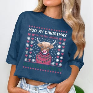 May include: A navy blue t-shirt with a festive design. The shirt features a Highland cow wearing a red sweater, surrounded by pink hearts and snowflakes. Text reads "Moo-ry Christmas Ya Filthy Animal."