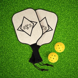 Könnte beinhalten: Zwei Pickleball-Schläger mit schwarzen Griffen und einem Monogramm-Design sowie zwei gelbe Pickleballs werden auf einer grünen Oberfläche gezeigt. Die Schläger haben eine hellfarbene Oberfläche mit schwarzem Rand und einem schwarzen rautenförmigen Design.