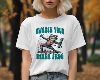 May include: White t-shirt with a green, blue, and red frog illustration. The frog is holding a rolled-up scroll and is surrounded by stars. The text "Awaken Your Inner Frog" is printed in green letters on the shirt.