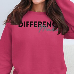 Op de afbeelding: Een magenta sweatshirt met de woorden "DIFFERENCE Made" in zwart en grijs op de borst gedrukt. De sweatshirt heeft lange mouwen en een ronde hals. Het algemene thema is vrijetijdskleding.