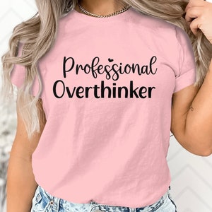 May include: A pale pink t-shirt featuring the black text "Professional Overthinker". The shirt has a crew neck and short sleeves. The text is in a cursive font and is centered on the front.
