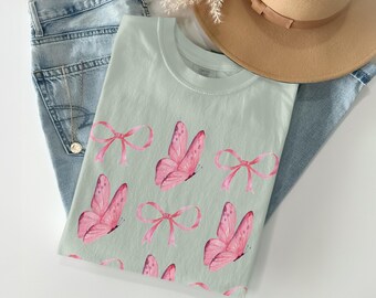 Peut inclure: Un t-shirt vert clair orné d'un motif de papillons et de nœuds roses. Le t-shirt est plié sur un jean bleu clair. Un chapeau beige est visible en arrière-plan.