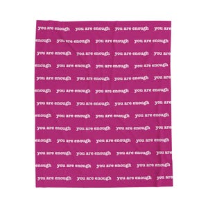 Può includere: Una coperta magenta con la scritta bianca "you are enough" ripetuta. Il testo è uniformemente distanziato, creando un motivo. La coperta, di forma rettangolare, sembra morbida e confortevole.