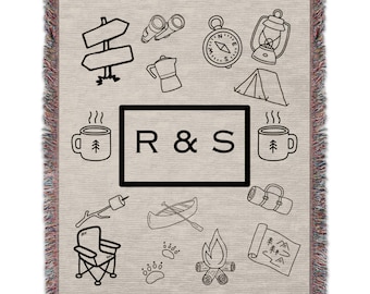 May include: A woven throw blanket with a beige background and black outlines of camping icons. The icons include a compass, a lantern, a tent, a canoe, a campfire, a mug, a bear paw print, a map, a chair, binoculars, a coffee pot, and a signpost. The blanket also features the text "R & S" in a black rectangle.