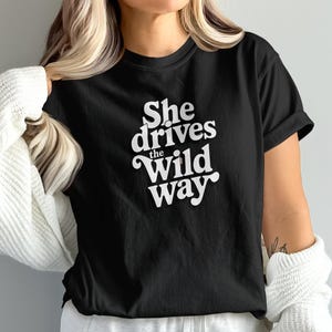Puede incluir: Camiseta negra con el texto blanco "She drives the Wild Way". La camiseta es de manga corta y tiene cuello redondo. La persona de la imagen lleva un suéter blanco sobre la camiseta.