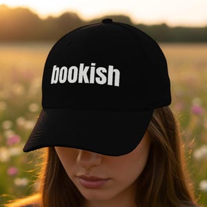 Op de afbeelding: Zwarte baseballpet met het witte woord "bookish" geborduurd op de voorkant. De pet heeft een gebogen klep en een knoop aan de bovenkant. De achtergrond is een wazig bloemenveld en zonlicht.
