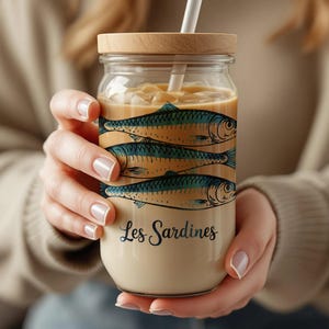 May include: A clear glass jar with a wooden lid and a straw, filled with a beverage. The jar is decorated with illustrations of sardines and the text "Les Sardines". The drink is light brown, and the jar is held by a person.