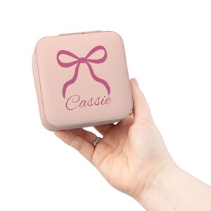 Peut inclure: Une boîte à bijoux carrée rose clair avec un motif de nœud et le nom "Cassie" en écriture cursive. La boîte est tenue dans une main sur fond blanc. Le nœud et le nom sont d'un rose plus foncé.