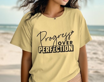 May include: A pale yellow short-sleeved t-shirt featuring the black text Progress OVER PERFECTION printed on the front. The word Progress is written in a cursive script font, while OVER PERFECTION is displayed in a bold, blocky, sans-serif typeface.