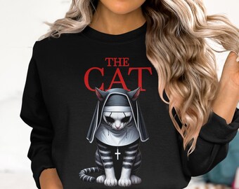 May include: Black sweatshirt featuring a graphic of a cat dressed as a nun, with the text "THE CAT" in red. The cat is black and white with a cross on its chest. The cat has black eye makeup.
