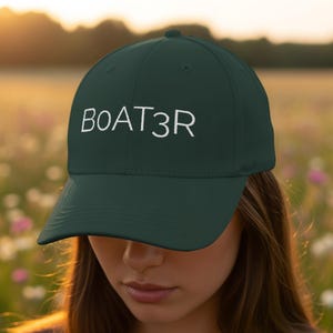 Puede incluir: Gorra de béisbol verde oscuro con la palabra "BOAT3R" bordada en blanco en la parte delantera. La gorra tiene una visera curva. El fondo es un campo de flores borroso.