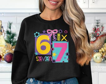 Peut inclure: Sweat-shirt noir avec un graphisme coloré représentant les chiffres "67" dans un design ludique. Le chiffre "6" est dans un encadré jaune avec une bordure violette, et le chiffre "7" est dans un encadré rose avec une bordure turquoise. Le mot "seven" est en dessous.