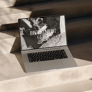 May include: A silver laptop computer with a black keyboard is open on a set of concrete steps. The laptop screen displays a black and white image of two people holding a glass chandelier with the text "INVESTMENT GUIDE" displayed on the screen.