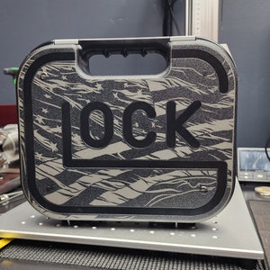 Könnte beinhalten: Ein schwarzer Kunststoff-Aufbewahrungskasten mit einem Griff und einem Tarnmuster. Der Kasten hat den Schriftzug "LOCK" auf der Vorderseite.