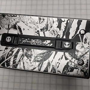 May include: A black metal ammo can with a graphic print of comic book characters, including Wolverine and Deadpool. The can has a black handle with a silver latch and a silver latch on the side. The text "LFG" is printed on the handle.