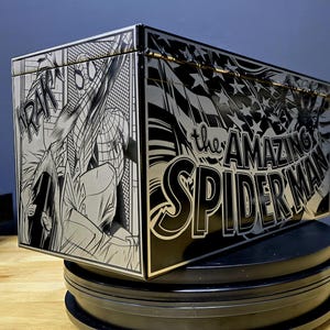 Op de afbeelding: Een zwarte metalen doos met een zilveren metallic afwerking. De doos heeft een stripboek-achtige illustratie van Spider-Man die door de lucht slingert. De tekst "the AMAZING SPIDER-MAN" is in vetgedrukte witte letters op de doos gedrukt.