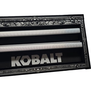 Puede incluir: Caja de herramientas negra con dos tiradores de plata y un logotipo "KOBALT" plateado en la parte delantera. La caja de herramientas tiene un borde plateado adornado con un diseño decorativo.
