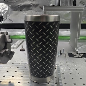 May include: A black and white stainless steel tumbler with a diamond plate pattern. The tumbler is sitting on a metal surface with a grid of holes.