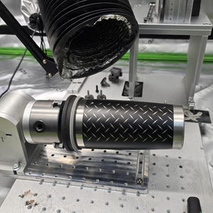 May include: A close-up of a black and silver industrial machine with a black and white patterned cylinder being held in place by a clamp. The cylinder is being inspected by a black bellows-like device.